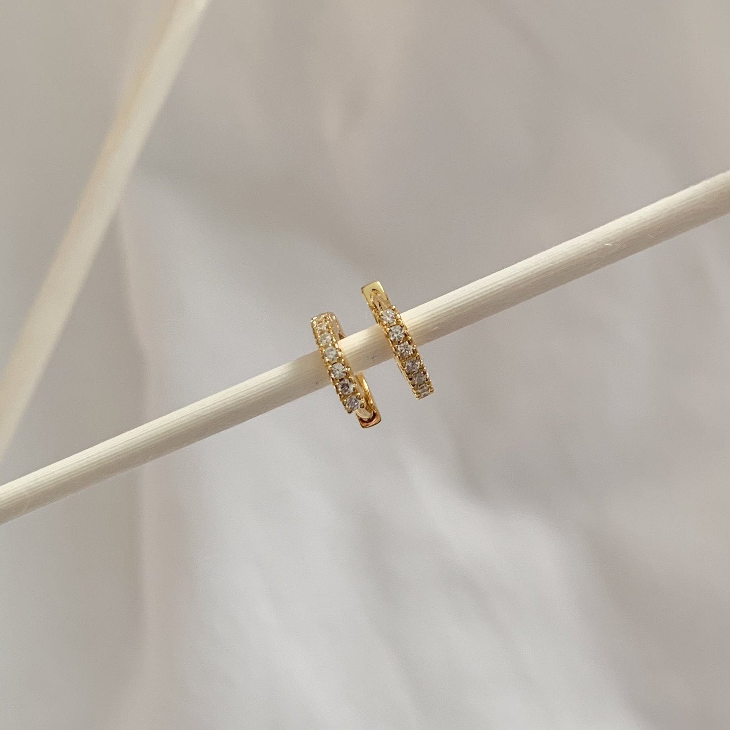 Gold hoop earrings with embedded stones on a light wooden stick against a neutral background