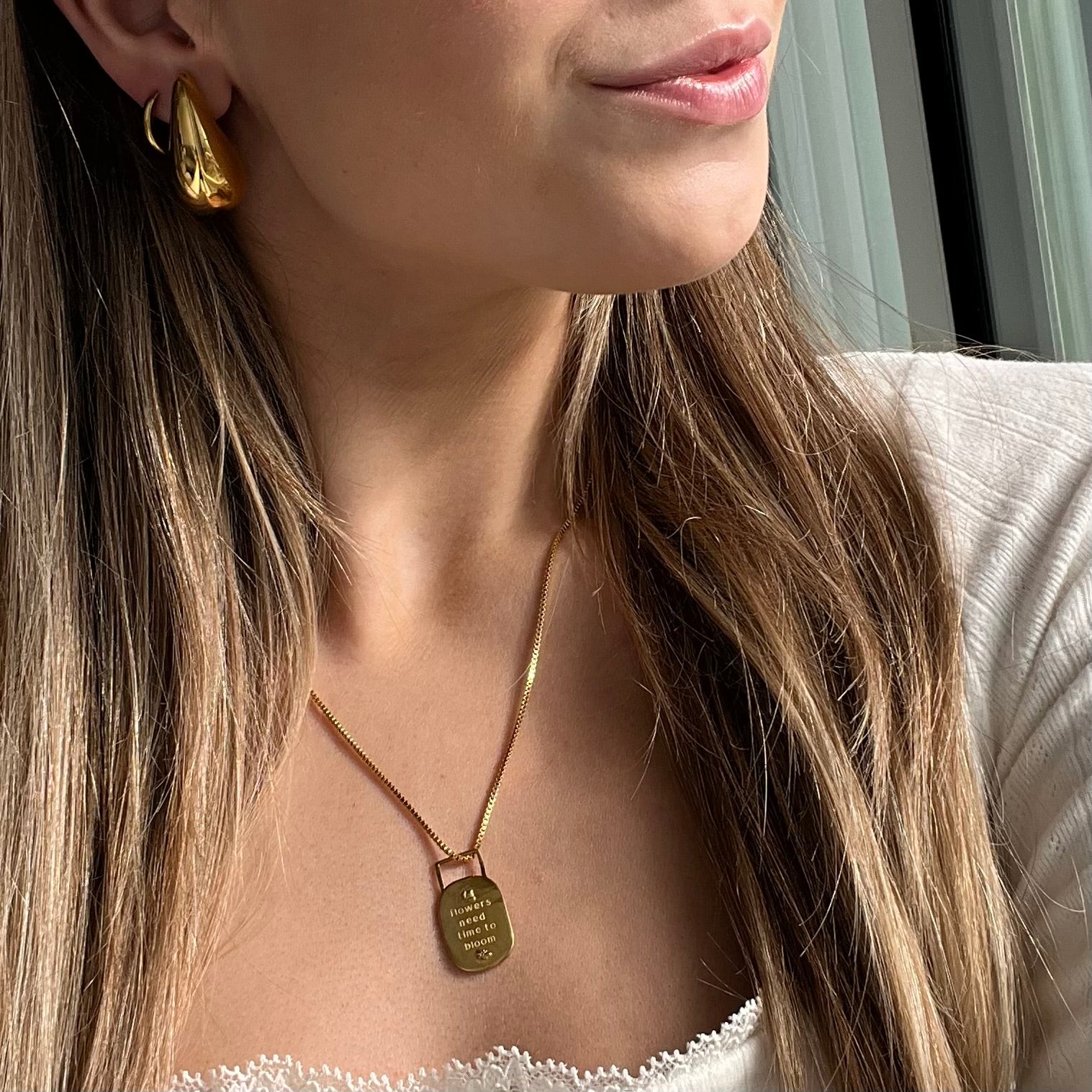 Close-up of a woman wearing a gold necklace with a pendant that says in writing "flowers need time to bloom"