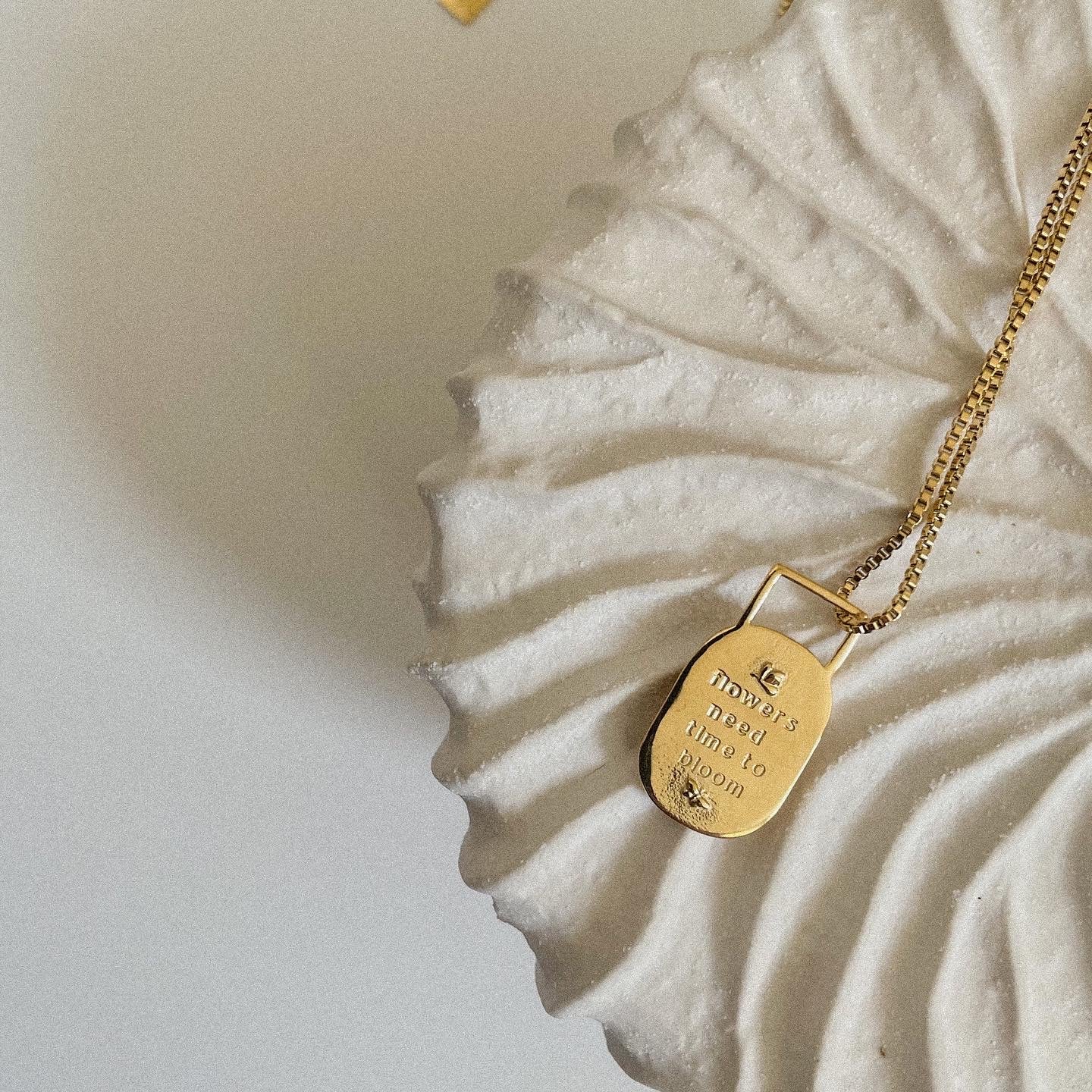 Gold necklace with a pendant that reads "flowers need time to bloom" on a textured white surface