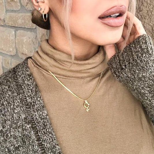 Woman wearing a gold necklace with a gold venus symbol pendant against a stone wall background
