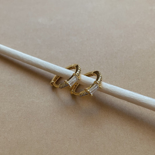 Gold hoop earrings with a pave crust and a rectangular white zirconia stone in the middle, on a beige background