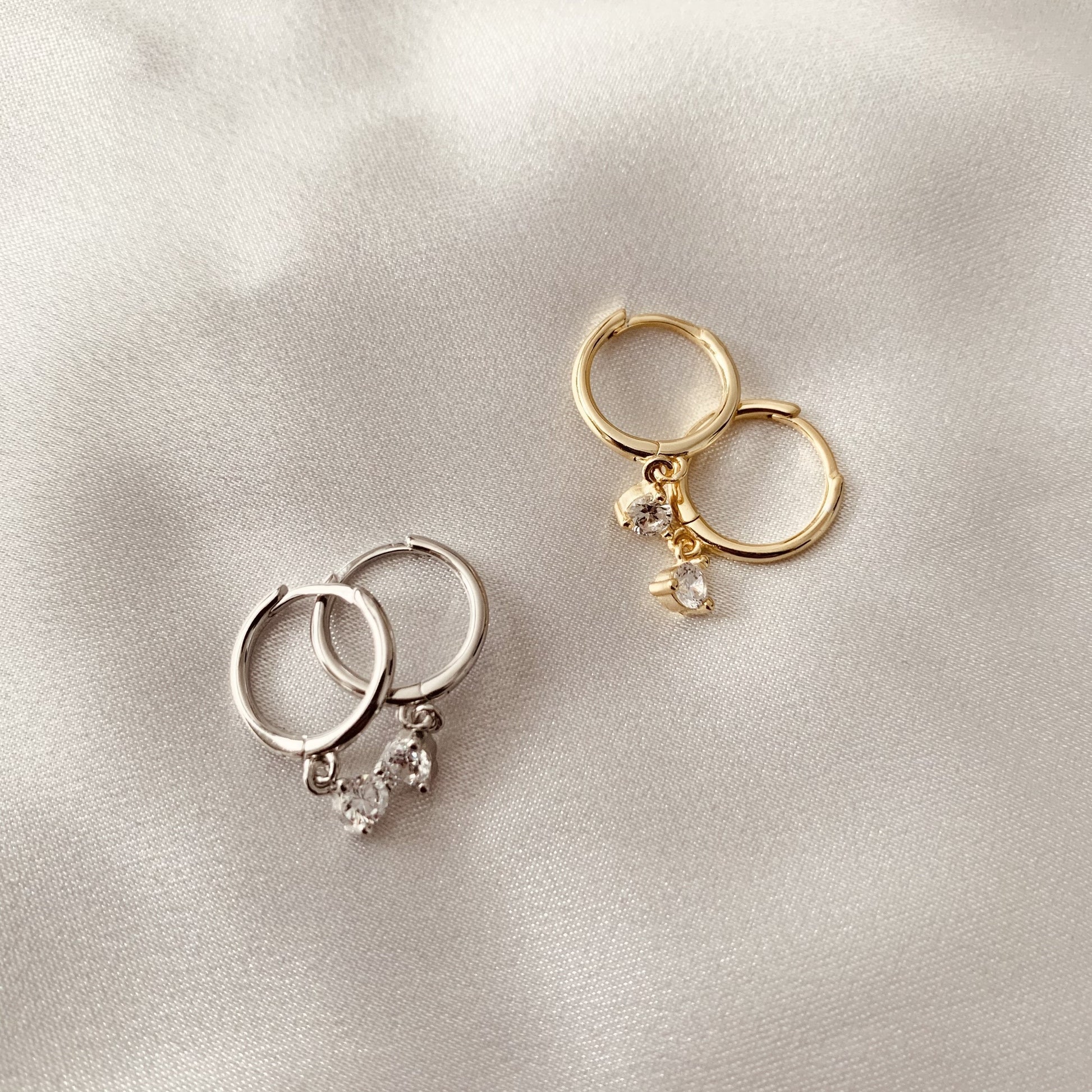 Two pairs of hoop earrings, one gold and one silver, with cubic zirconia drop, on a light gray background.