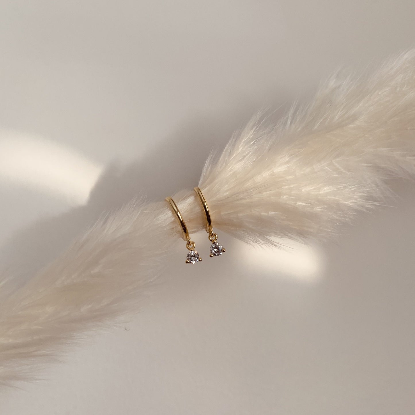 Gold hoop earrings with small dangling white zirconia charms on a white feather surface