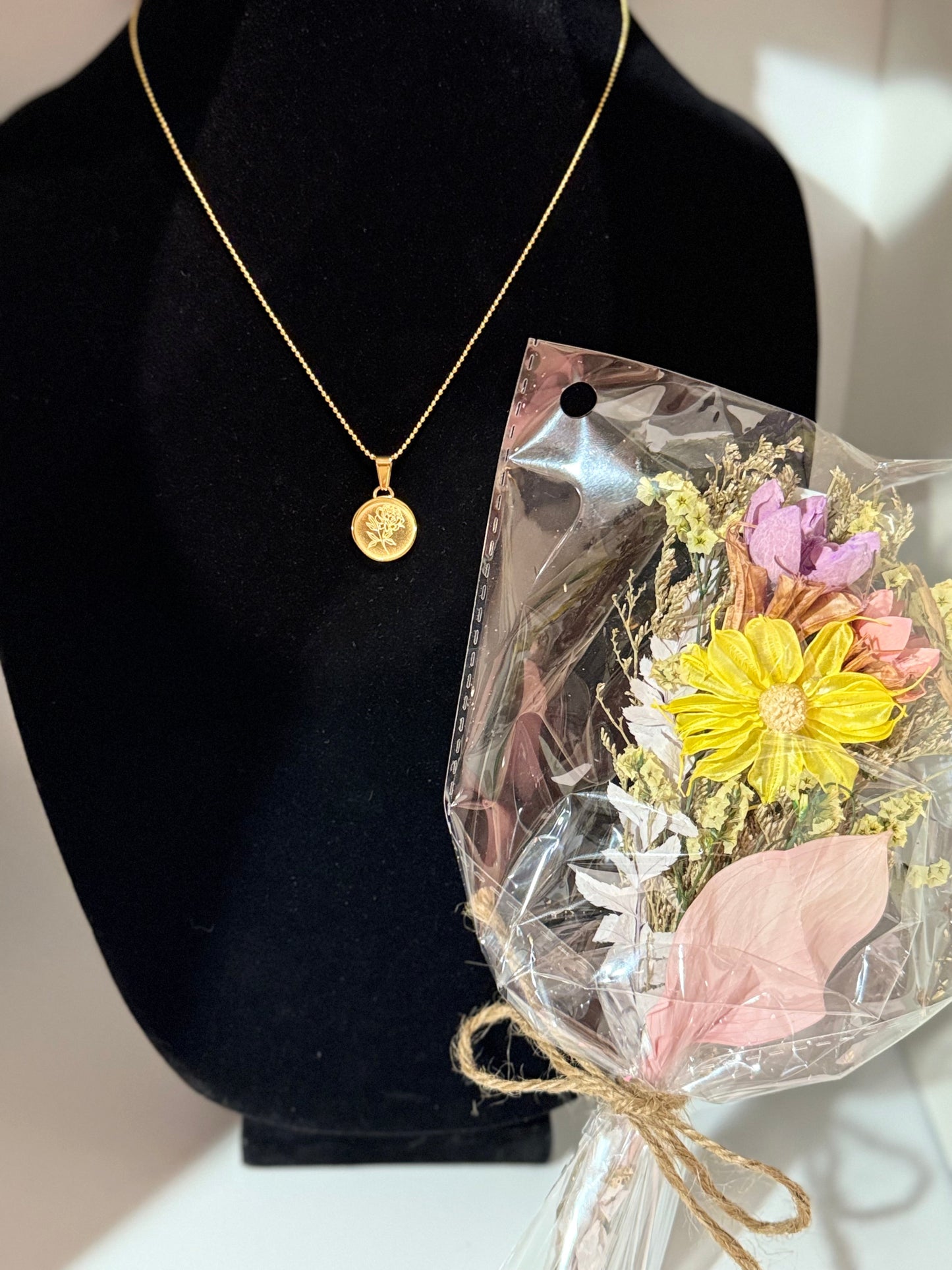 Gold pendant necklace with a floral design on a black stand with a bouquet of flowers in clear packaging.