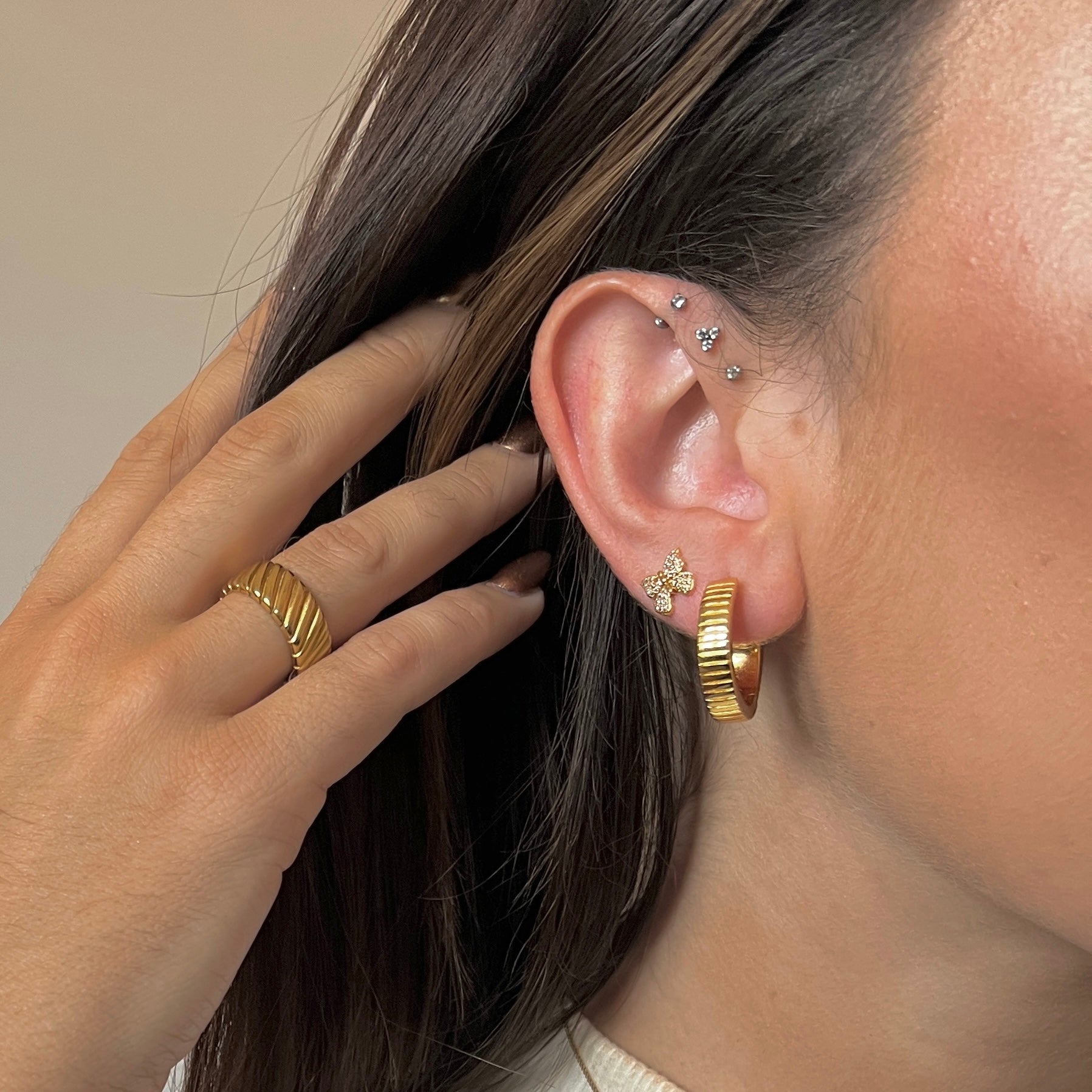 Close-up of an ear with gold hoop earrings and a ring, with a neutral background