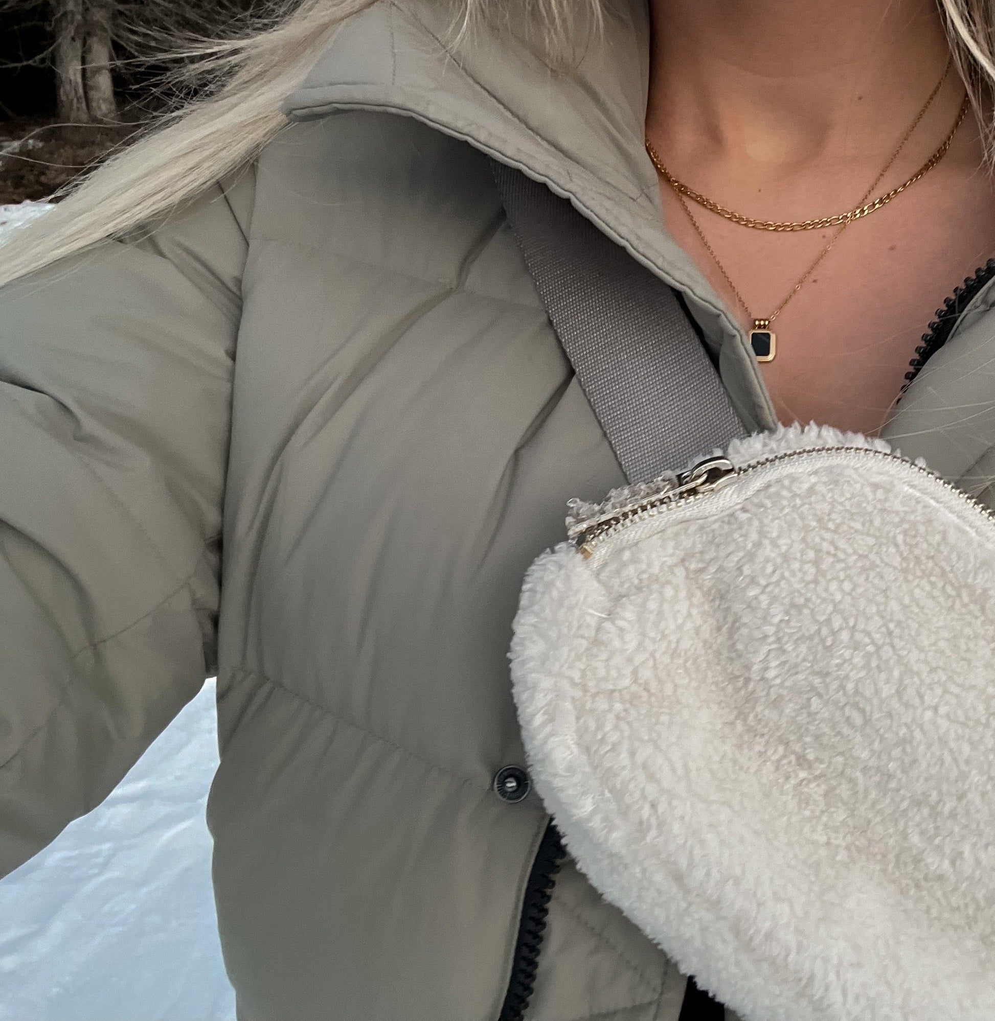Person wearing a light gray puffer jacket with a focus on her 2 layered gold necklace with a square gold pendant with a black stone in the middle.