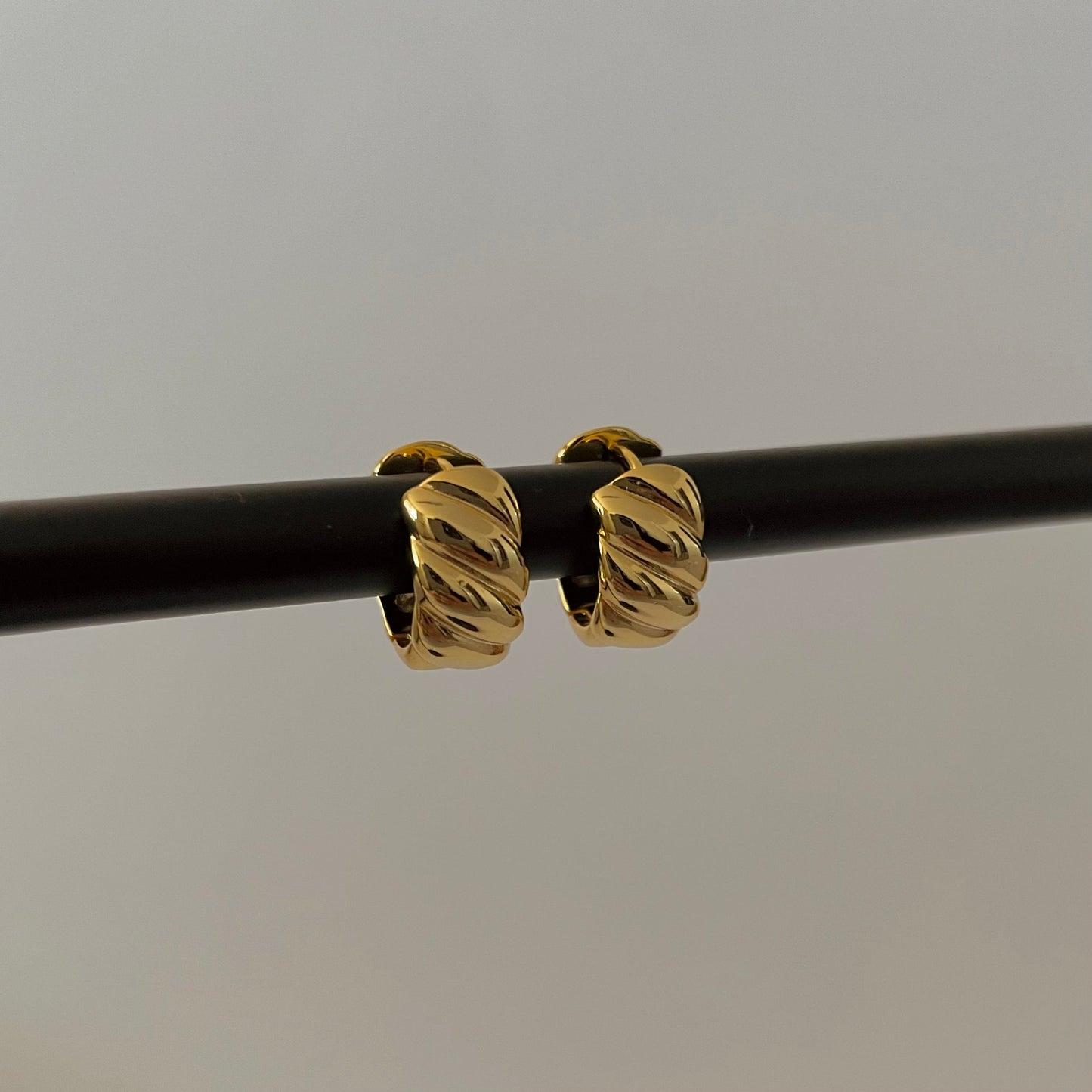 gold sculptural hoop huggies on a black rod, plain grey background