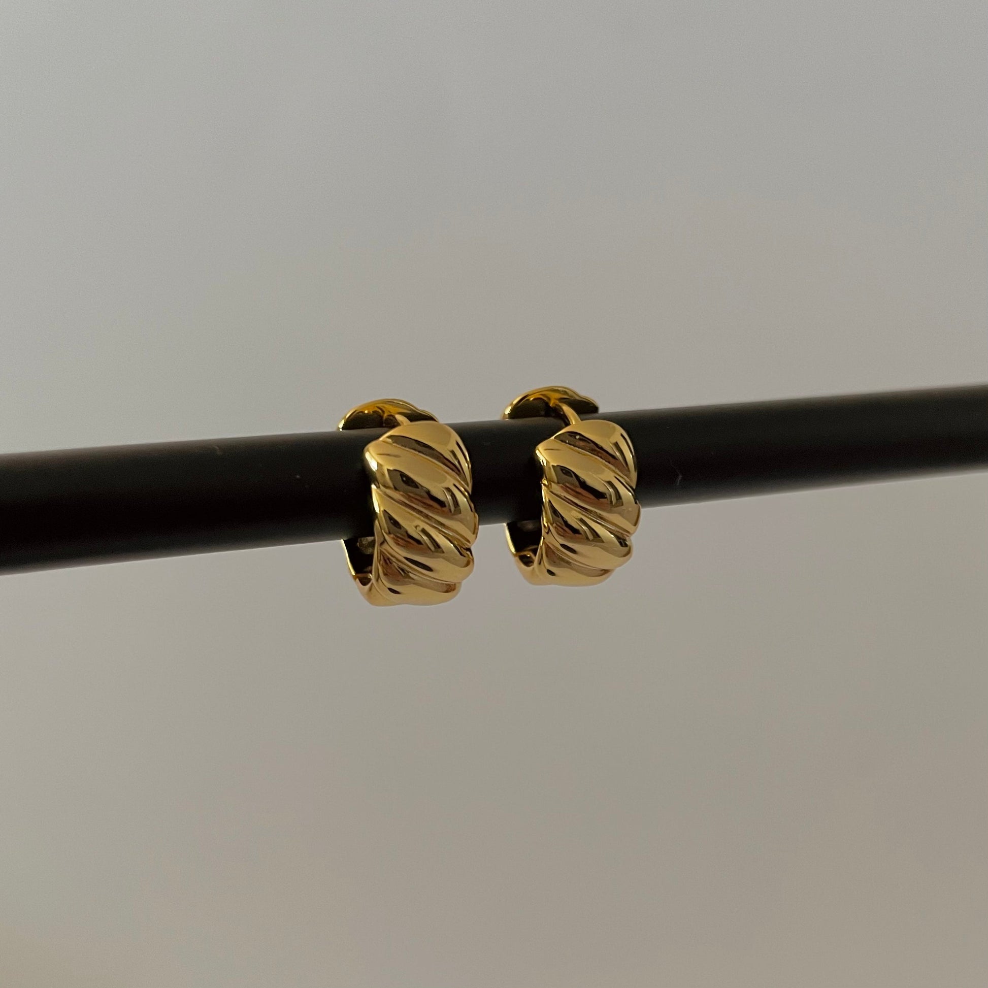 gold sculptural hoop huggies on a black rod, plain grey background