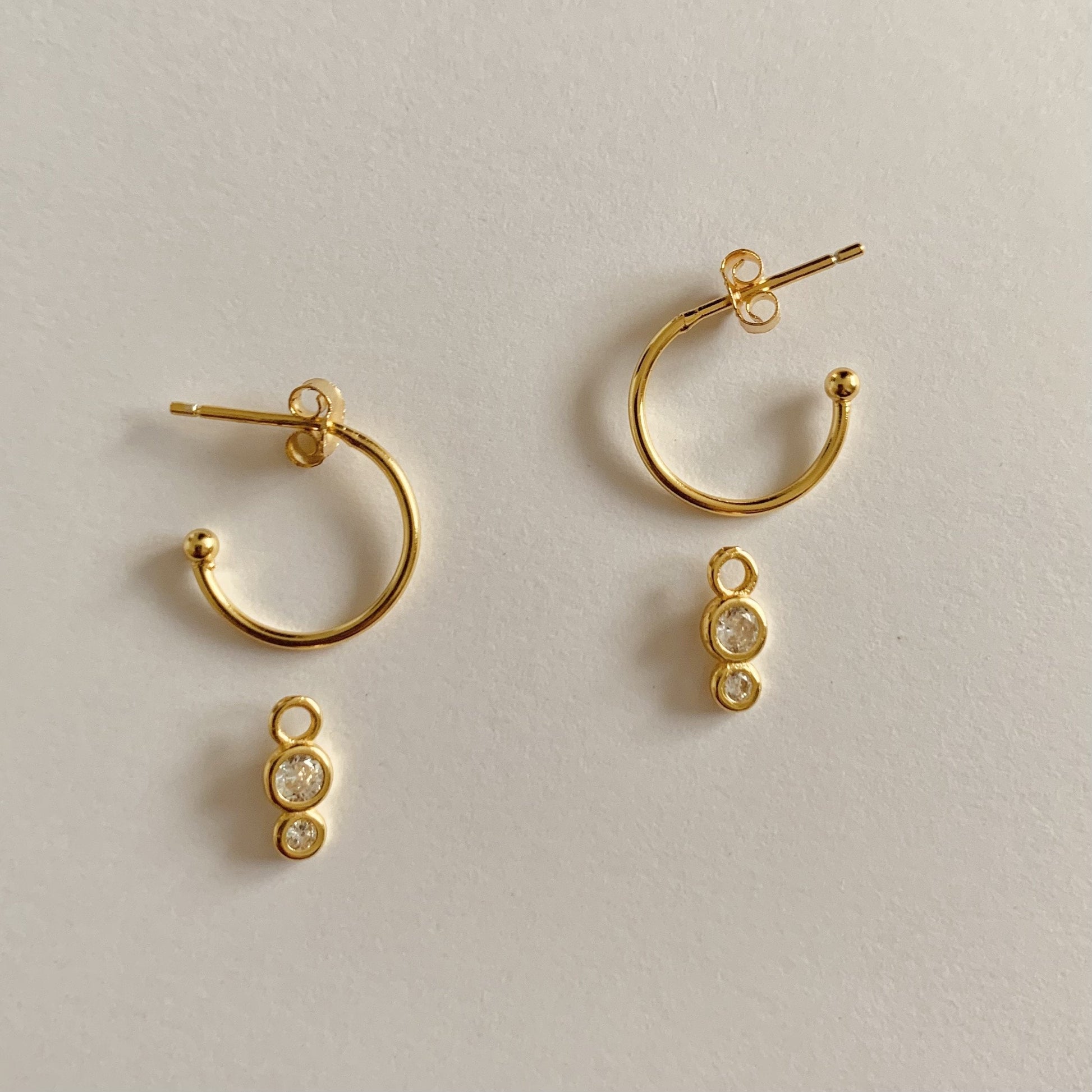 Set of gold hoop earrings with small clear dangling crystal spheres, top sphere larger and bottom sphere, on a plain paper textured background.
