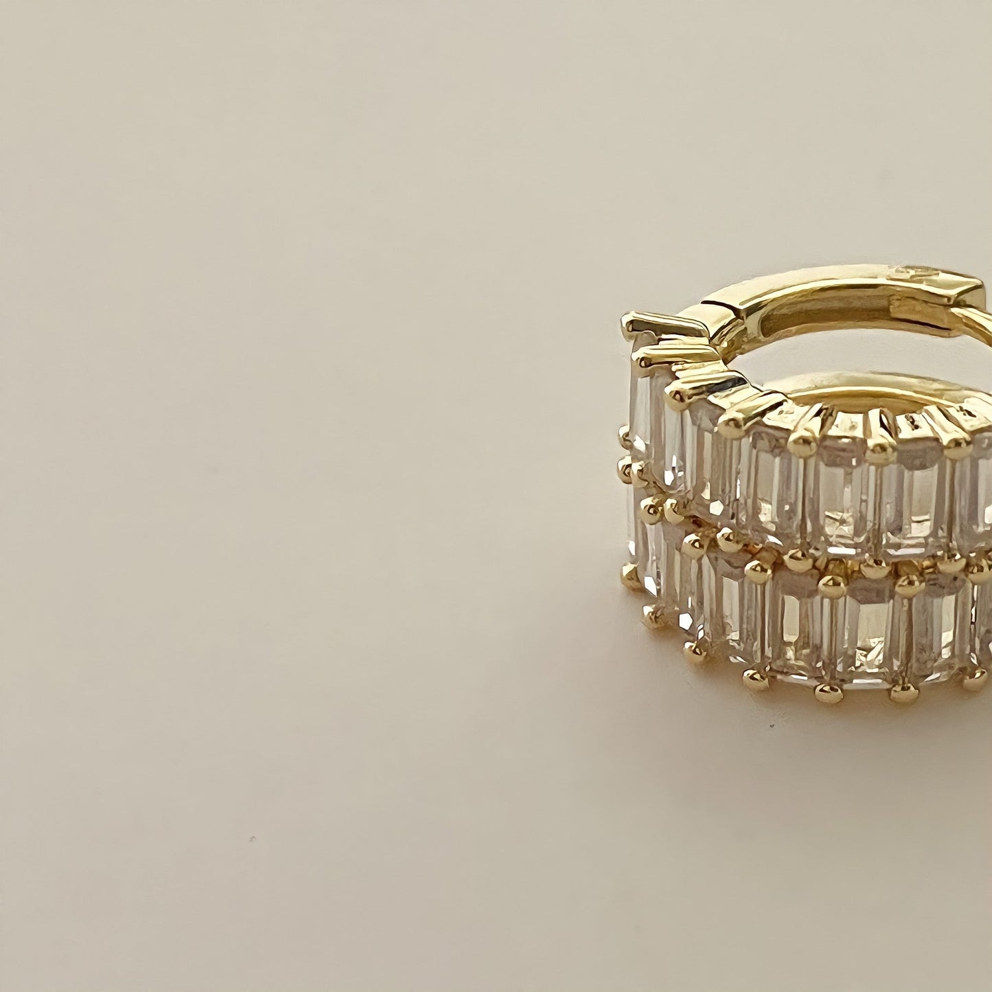 close up stacked gold earrings crusted with clear rectangular gemstones, on a beige background.