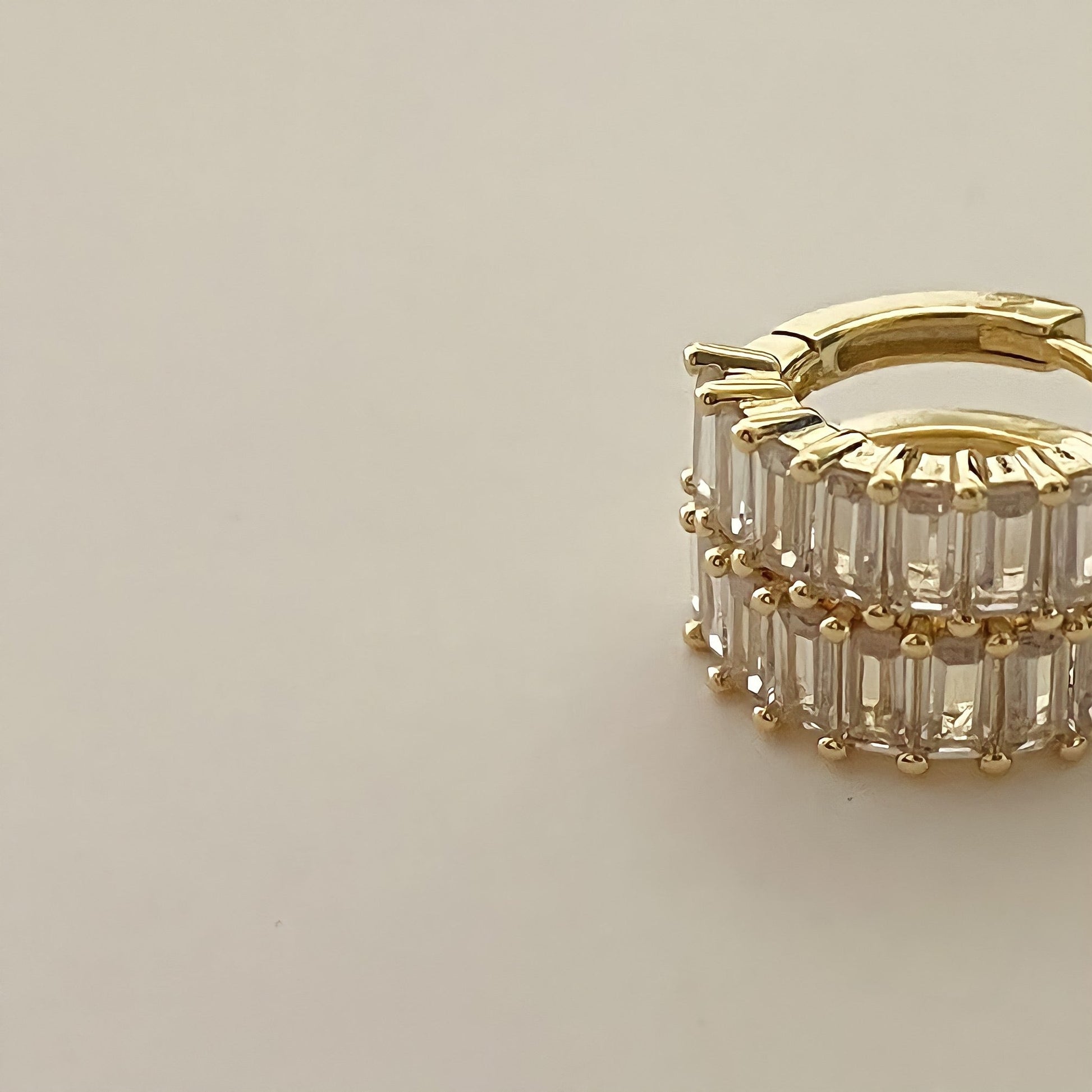 close up stacked gold earrings crusted with clear rectangular gemstones, on a beige background.