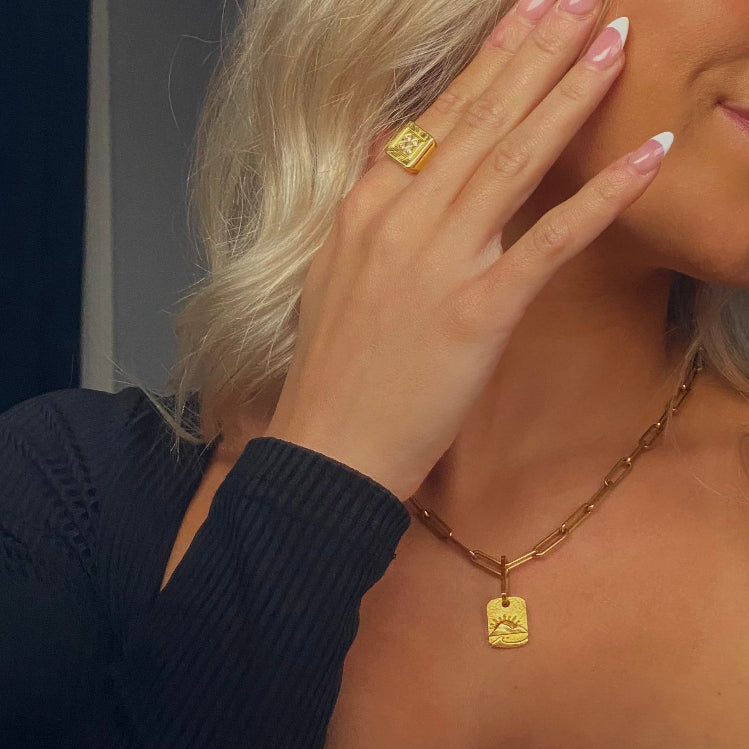 Woman wearing a gold sunset pendant necklace and ring, with a neutral background