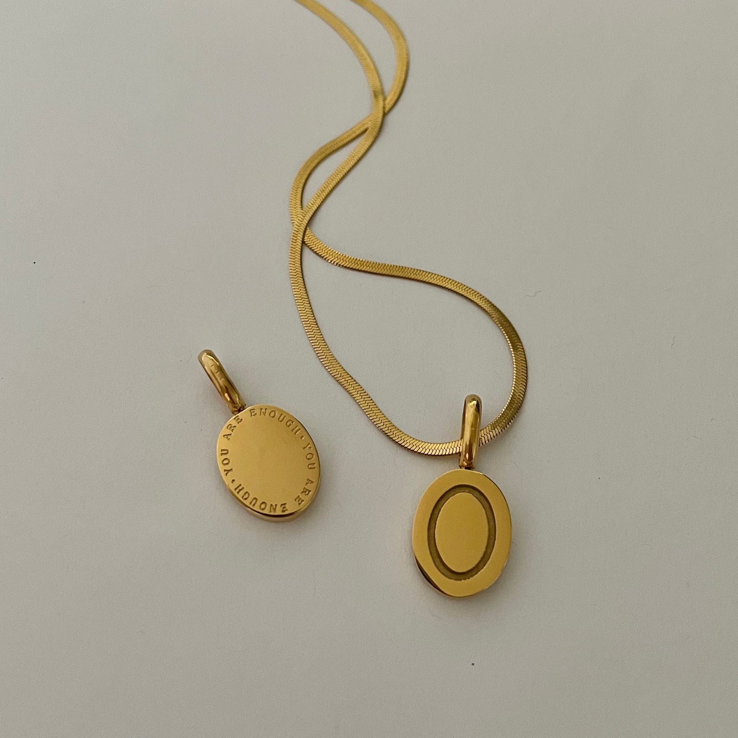 Gold necklace with an oval pendant that reads "you are enough" on a light gray background