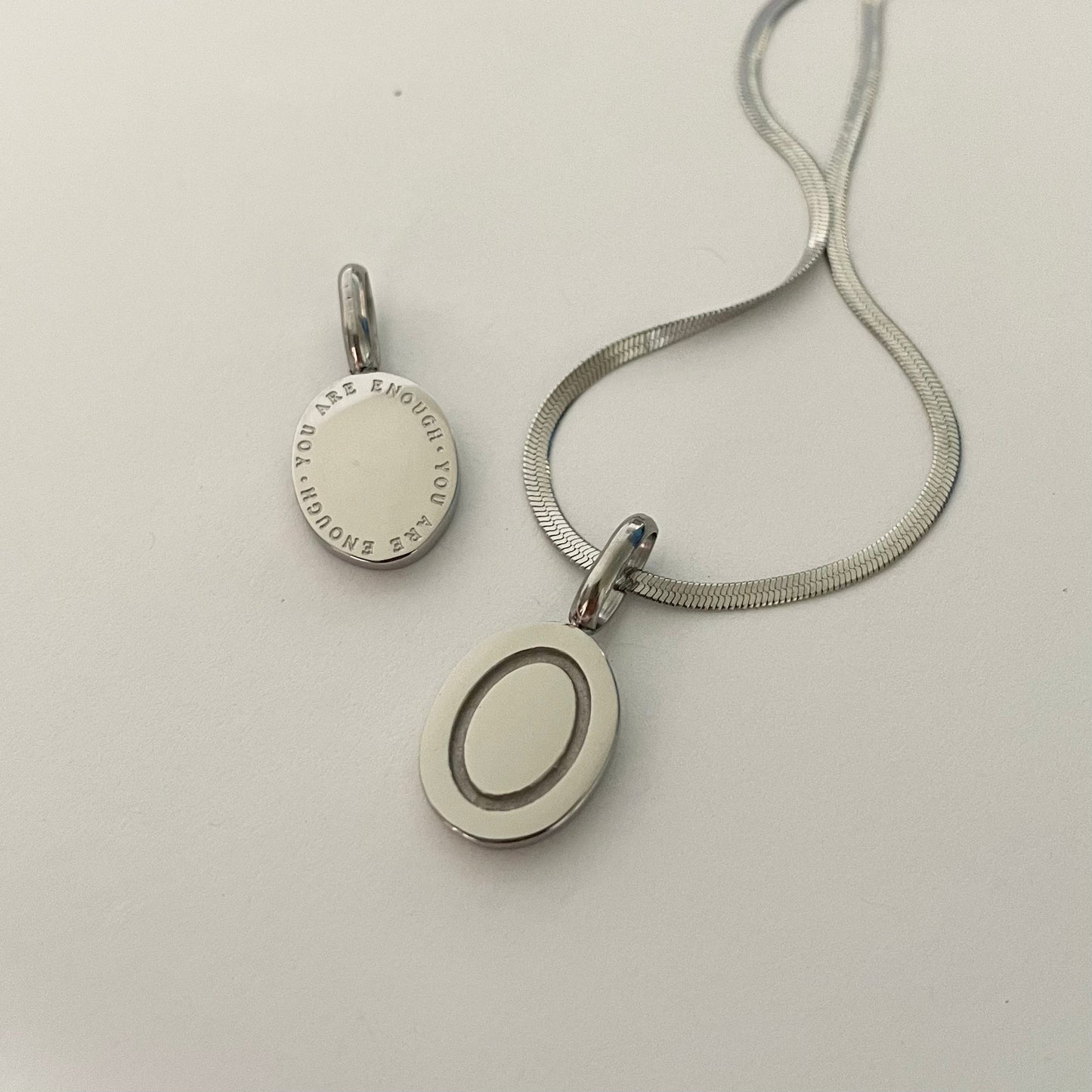 Silver necklace with a round pendant that reads "you are enough" attattched to a silver herringbone necklace  on a light gray background