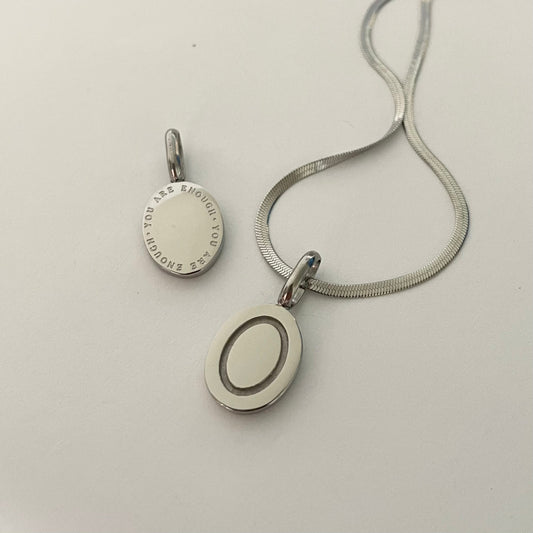 Silver necklace with a round pendant that reads "you are enough" attattched to a silver herringbone necklace  on a light gray background